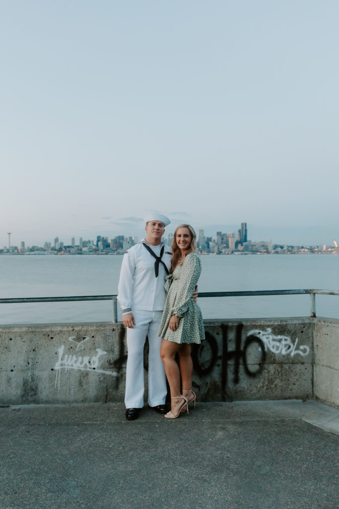 A couple standing next to each other smiling with the seattle skyline sprawled out behind them during their washington engagement photos