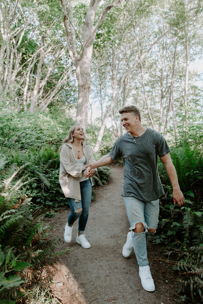 A couple holding hands and laughing as they walk down a path in a park during their Seattle engagement photos