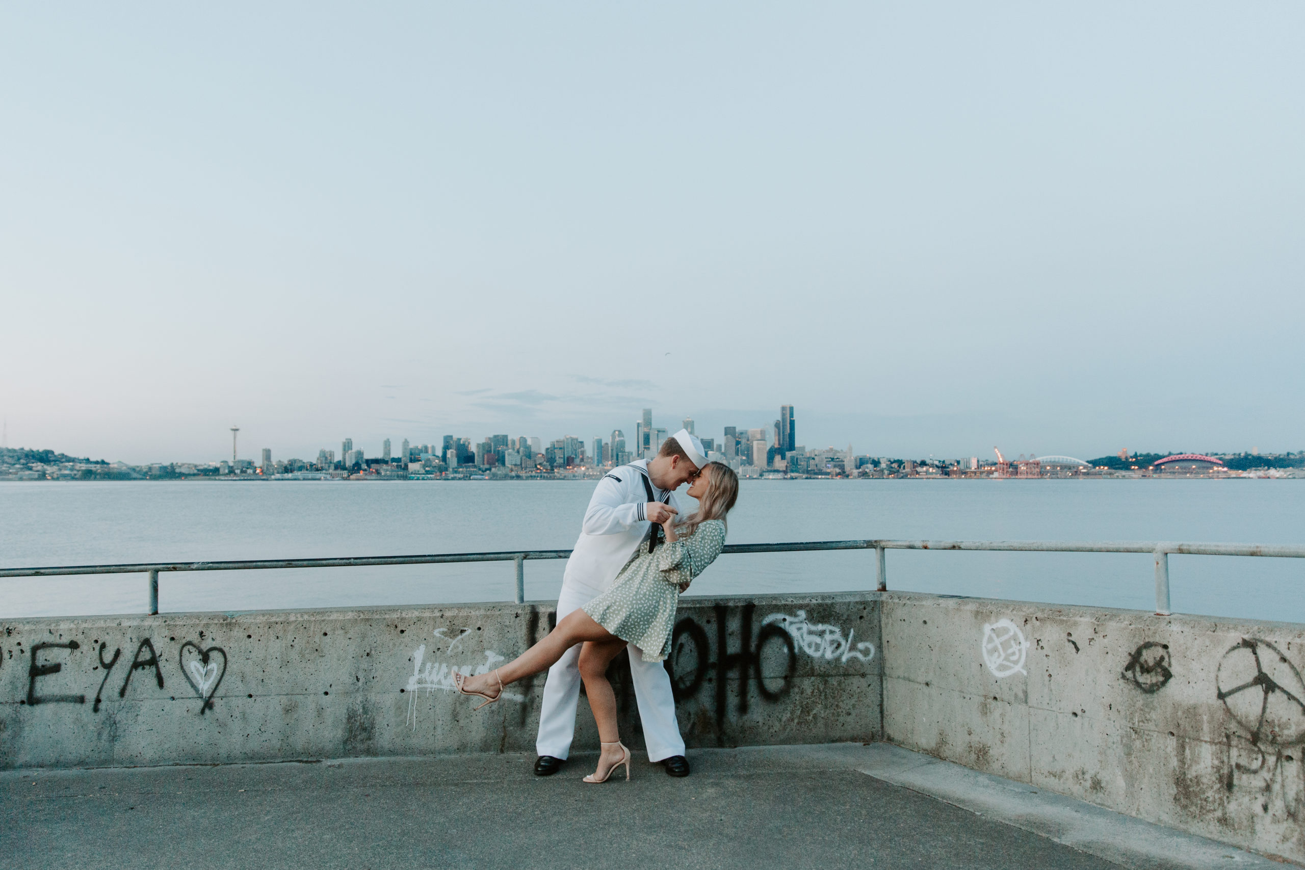 A couple standing on the edge of the water with the seattle skyline in the background as the man is dipping his partner and giving her a kiss during their Washington engagement photos