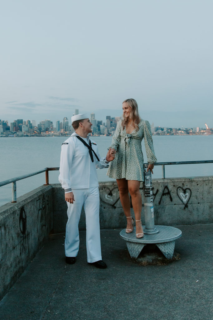 A woman standing up on a elevated surface while her partner is standing on the ground holding her hand and they are looking at each other with the Seattle skyline in the back during their summer engagement photos