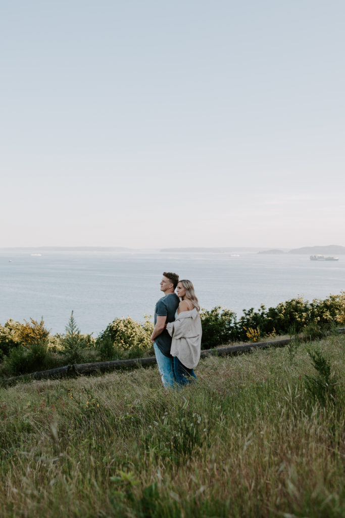 A couple standing in the middle of a field with the woman standing behind her partner with water in the background during their Discovery Park engagement photos in Seattle