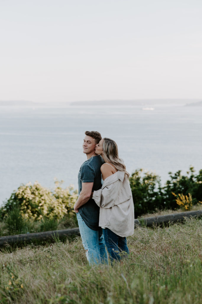 A woman standing behind her partner with her arms around him as she is giving him a kiss on the cheek during their Elliott Bay engagement photos