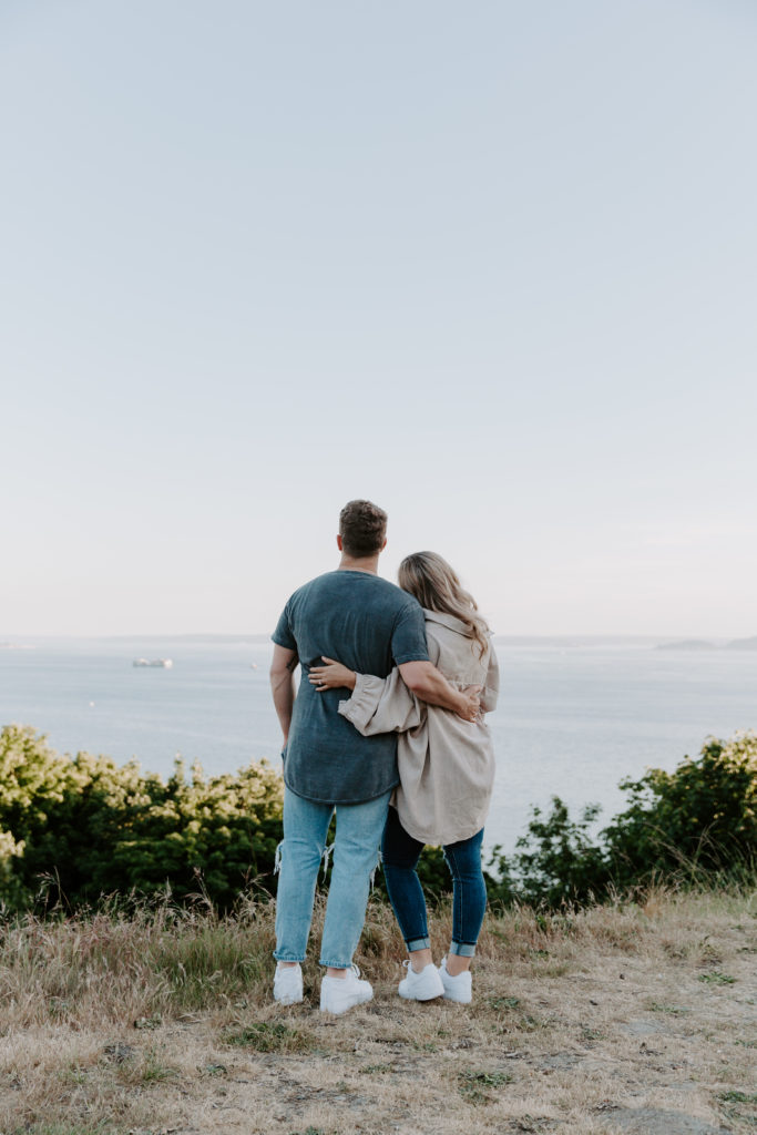 A couple looking off over Elliott Bay with their arms around each other during their Discovery Park Engagement photos