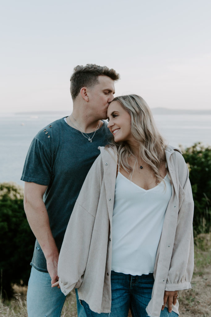 A man kissing his partner on the forehead as she is smiling and as they are holding hands during their summer Seattle engagement photos