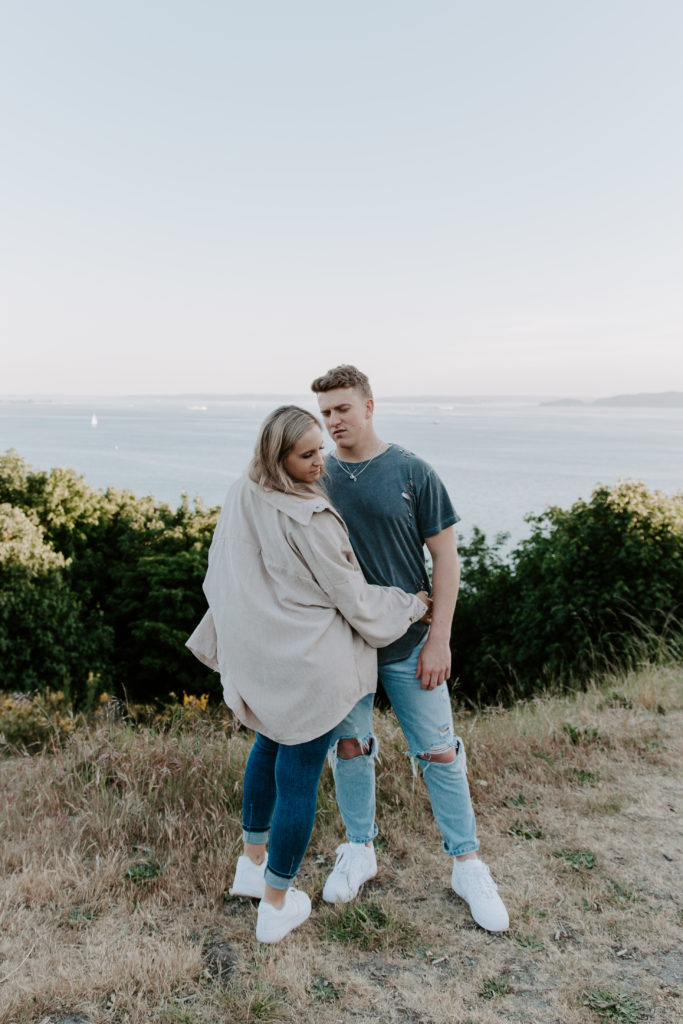 A man looking down at his partner as she is holding on to him and looking down at the ground during their Elliot Bay engagement photos