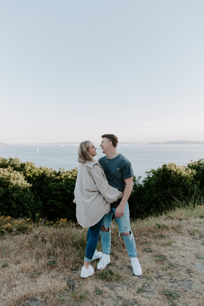 A woman holding on to her partners hip as she looks up at him and he is looking off into the distance during their Discovery Park engagement photos in Seattle