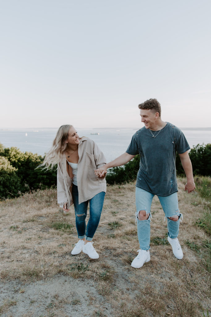 A couple holding hands and walking away from the bay laughing during their summer engagement photos in Seattle