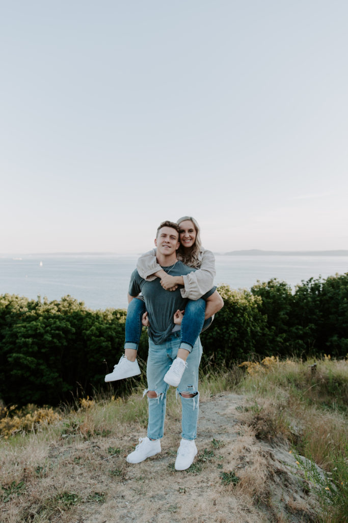 A man giving his partner a piggy back ride as they are both smiling and Elliott Bay in the background