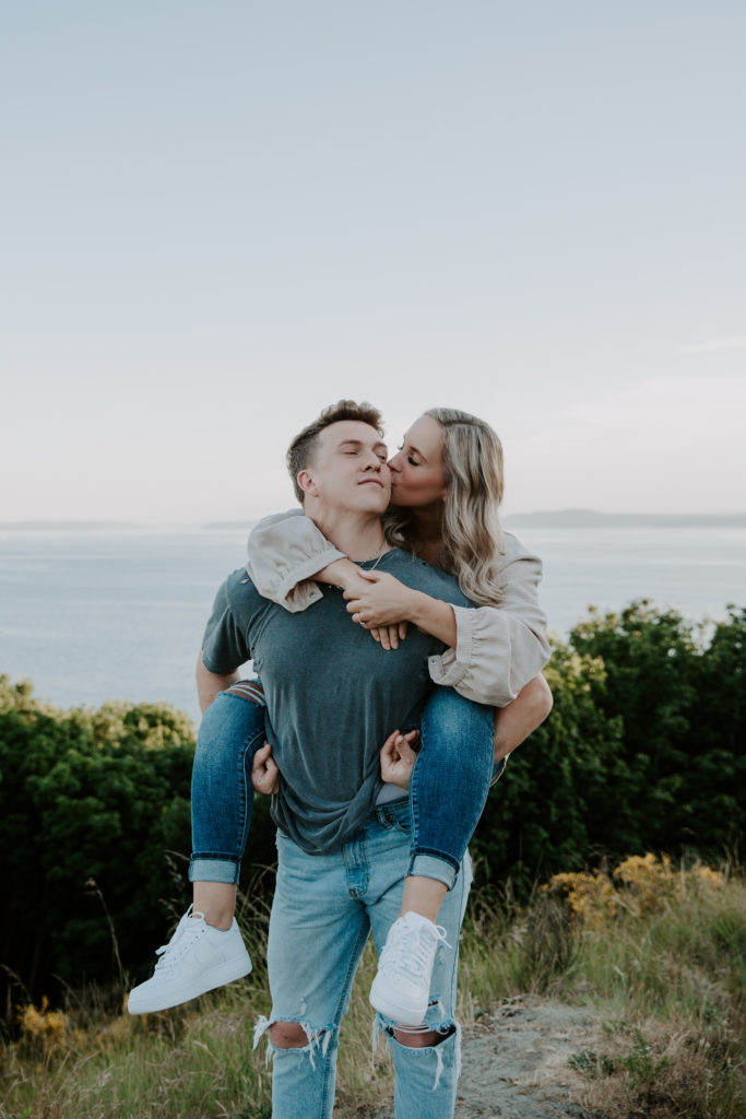 A woman kissing her partner on the cheek while he is giving her a piggy back ride during their Discovery Park engagement photos