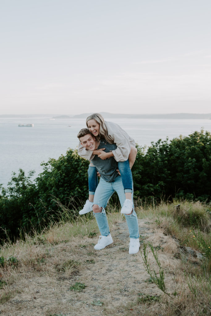 A man giving his new fiance a piggy back ride as they are both laughing and Elliott Bay behind them