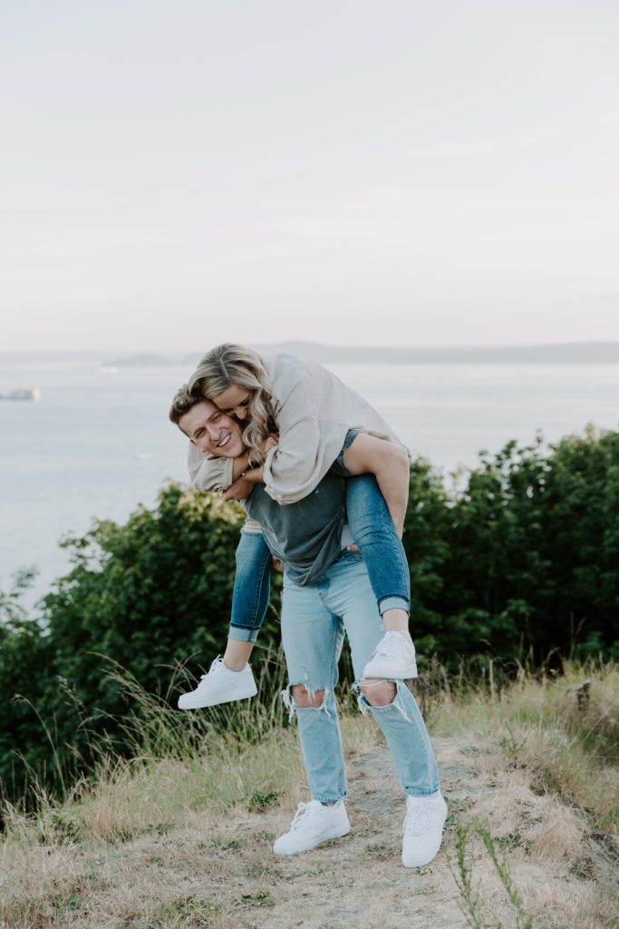 A man giving his partner a piggy back ride as she holds on to him really tightly and they are smiling during their Discovery Park engagement photos in Washington