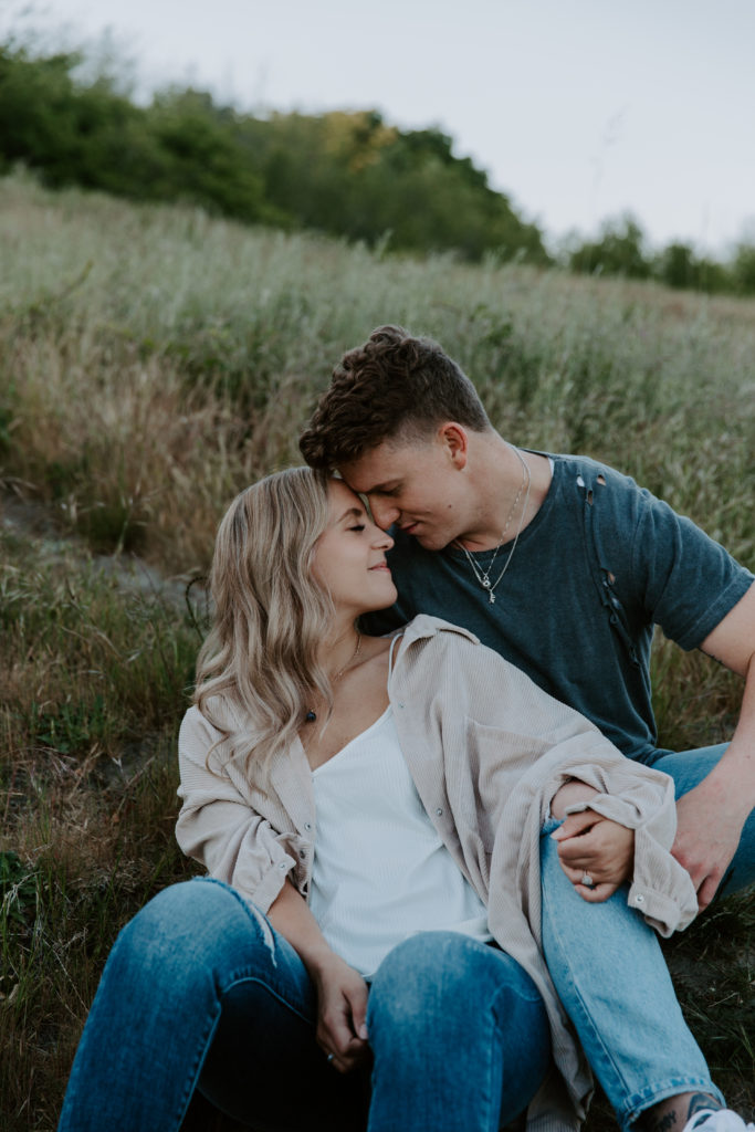 A couple sitting in a field with their foreheads leaning against the others and holding hands during their summer engagement photos in Seattle