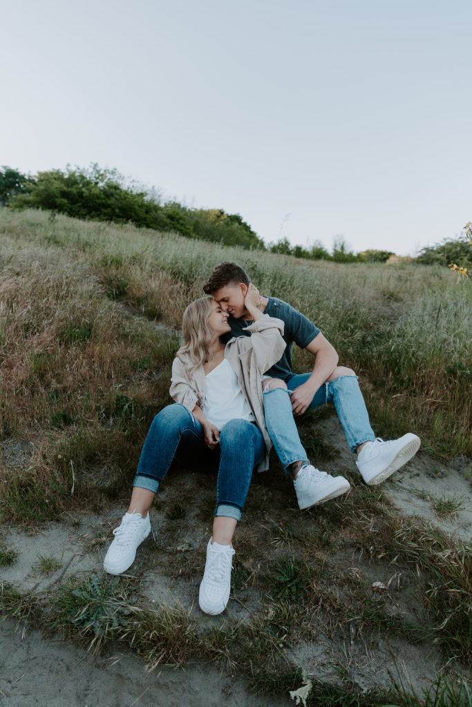 A couple sitting on the ground surrounded by tall grass as the woman is holding the mans neck and bringing him in to her during their Discovery Park engagement photos in Washington