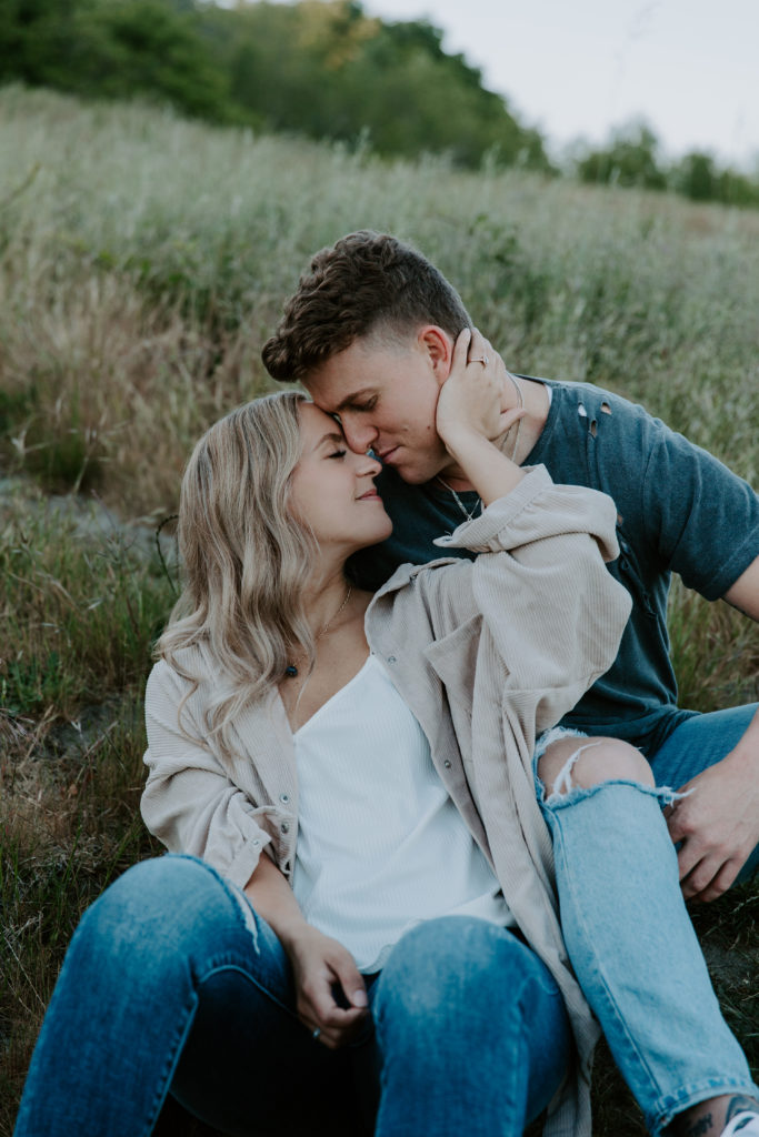 A couple sitting on the ground surrounded by tall grass as the woman is holding the mans neck and bringing him in to her during their Discovery Park engagement photos in Seattle