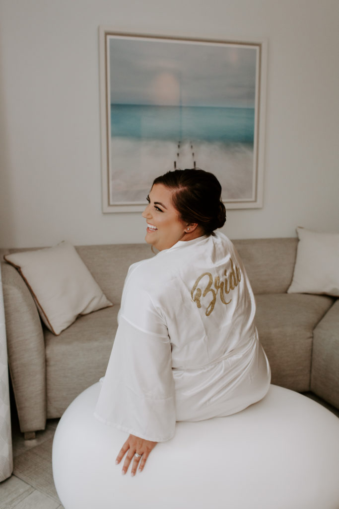 The bride sitting on an ottoman laughing over her shoulder before getting her wedding dress on