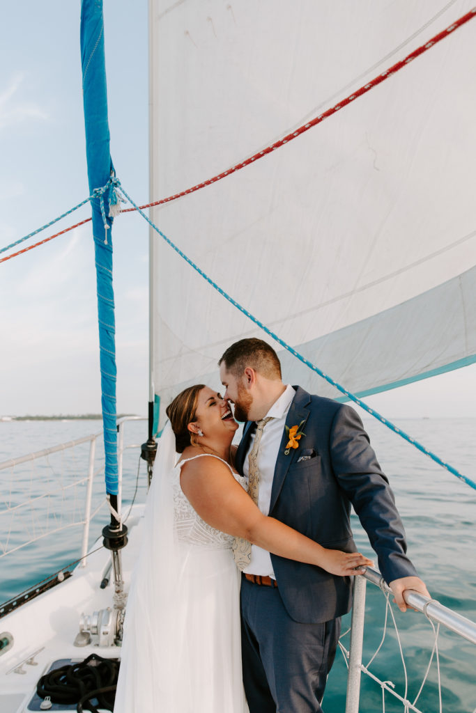 A couple in wedding attire leaning up against the railing of a sail boat while the woman is looking at her partner and laughing during their key west summer elopement