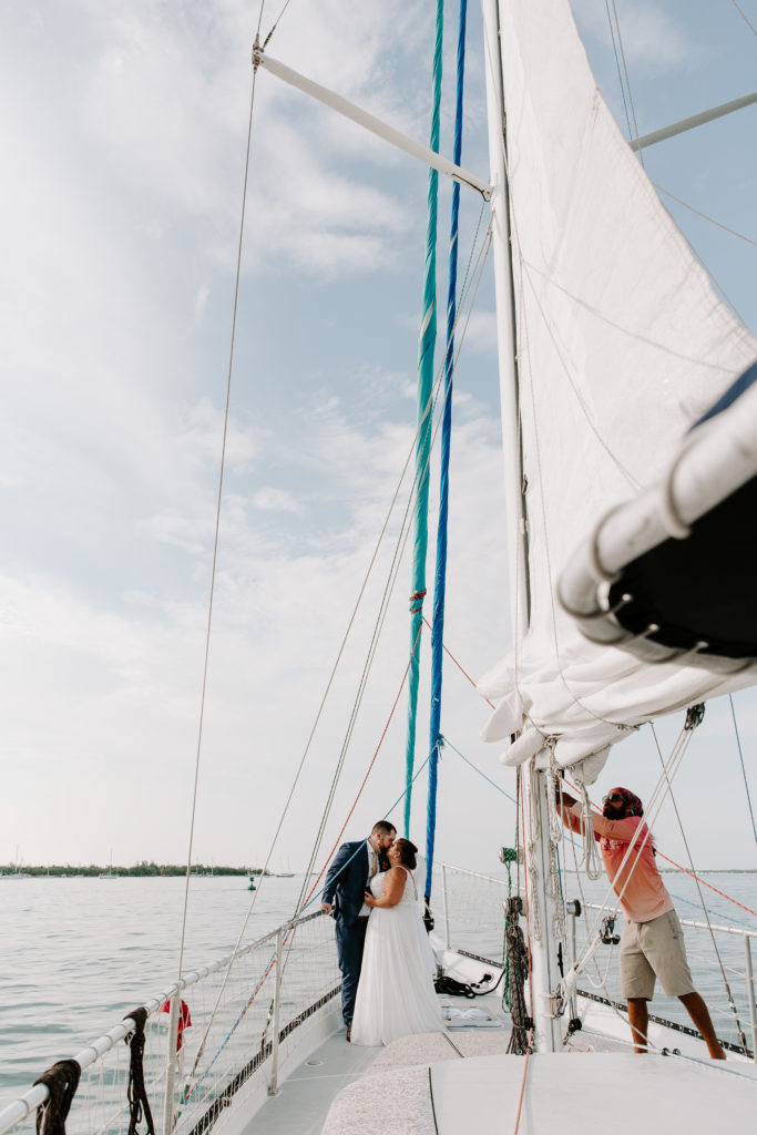 A couple standing at the front of a sail boat sharing a kiss as one of the crew members gets the sails up during their key west elopement