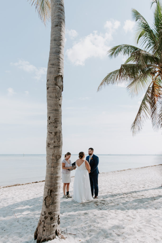 A couple and their officiant standing between palm trees on Smathers beach for their ceremony