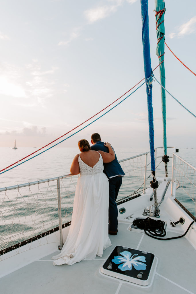 A couple standing at the edge of a sail boat leaning against the railing as the woman has her arm wrapped over her partners back admiring the sunset at the end of their all day florida elopement