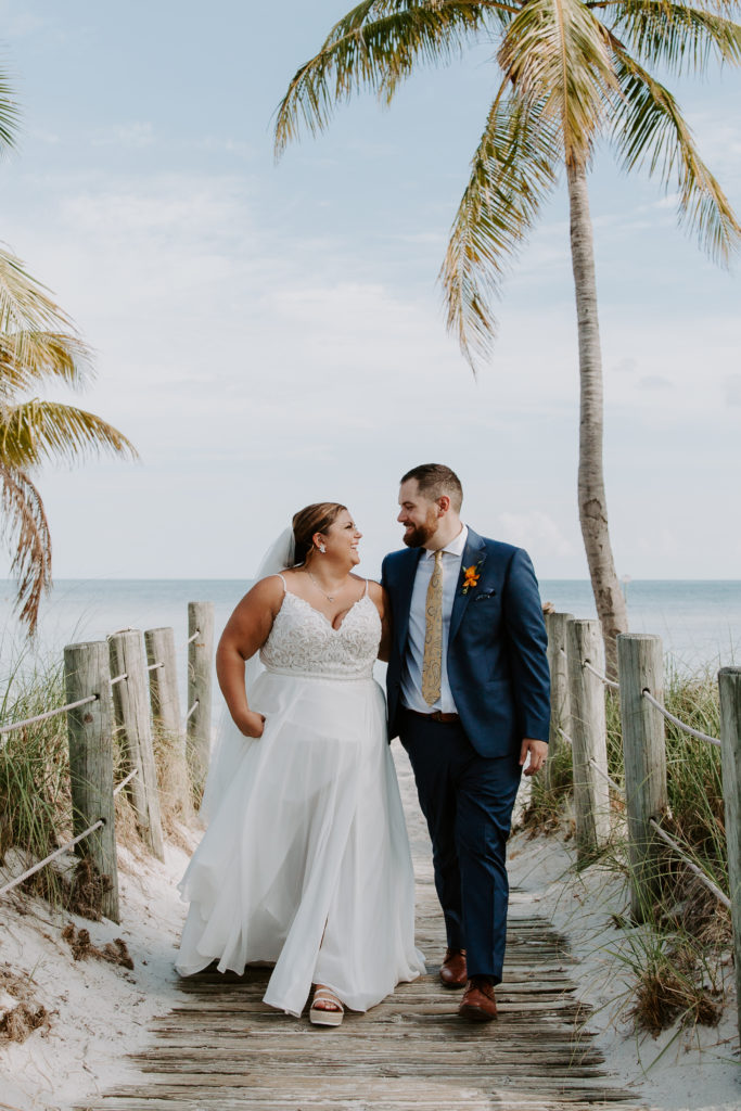 A couple in wedding attire holding hands and laughing at each other as they walk down the boardwalk on Smathers beach in Key West