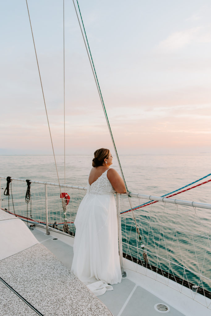 A woman in a wedding dress standing on the edge of a sail boat looking off at the sunset during their sunset cruise at the end of the florida elopement day