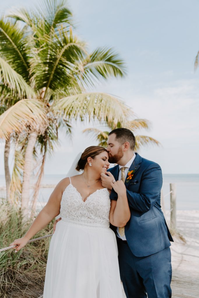 A man and a woman standing on a beach boardwalk with palm trees as the man kisses his partner on the forehead during their florida keys elopement
