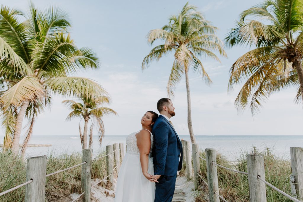 A couple standing back to back holding hands with the woman leaning her hear back on her partner during their all day beach town elopement