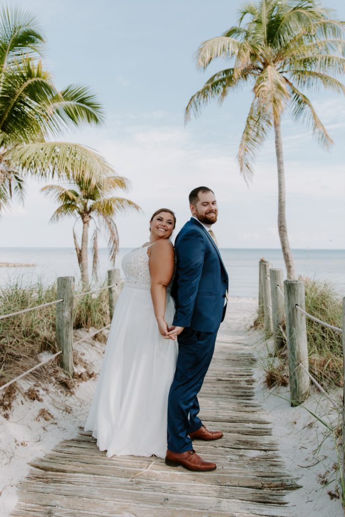 A newly wed couple standing back to back holding hands on a beach boardwalk with palm trees around them during their key west elopement