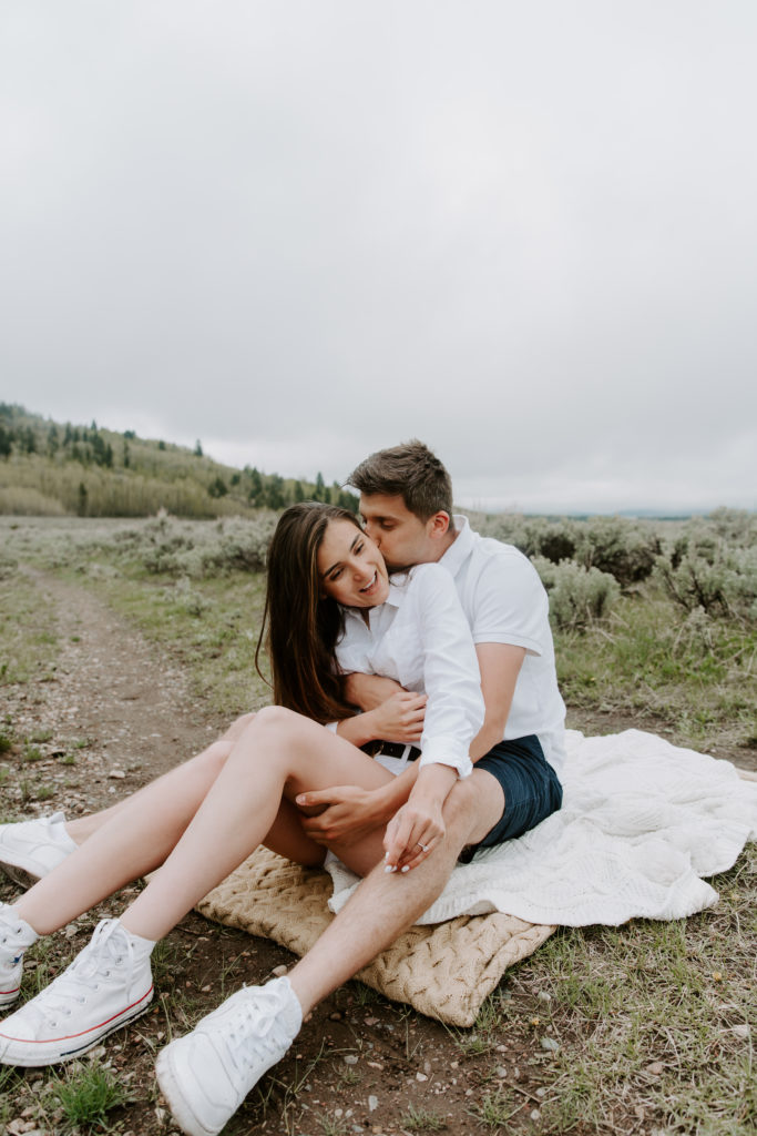 A man giving his partner a kiss on the cheek as she is smiling and they are both sitting on a blanket outside of Grand Teton National Park