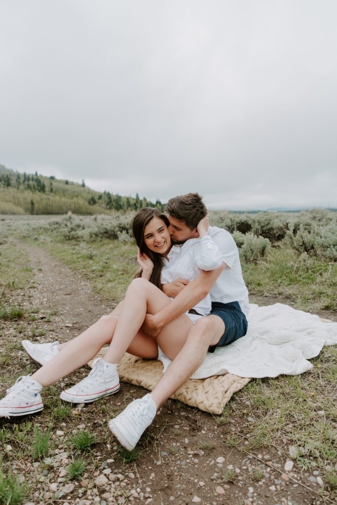 A man giving his partner a kiss on the cheek as she is holding her hair back and sitting in his lap during their Jackson Hole couple photos