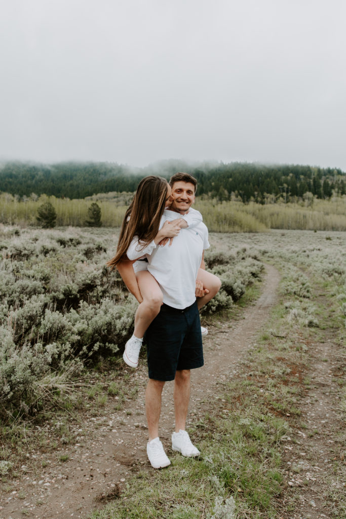 A woman giving her partner a kiss on the cheek as he is giving her a piggy back ride during their Wyoming engagement photos in Jackson Hole