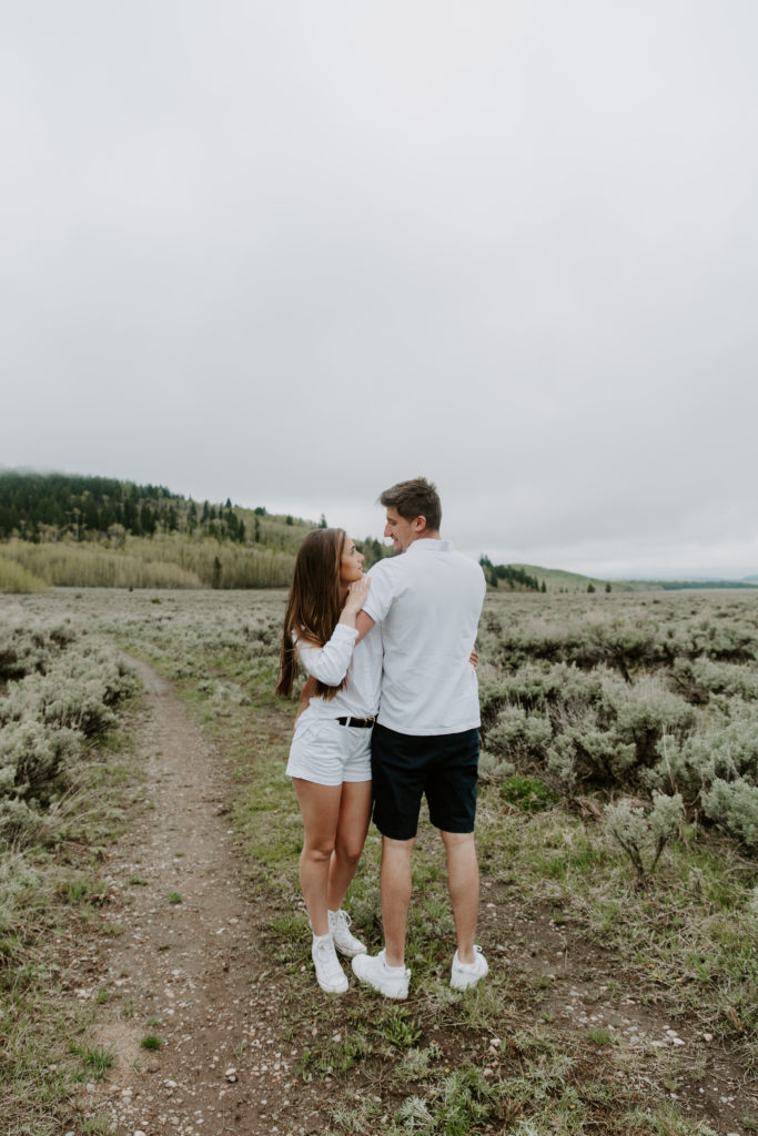 A couple standing in a field with the mountains behind them during their engagement photos outside of Grand Teton National Park
