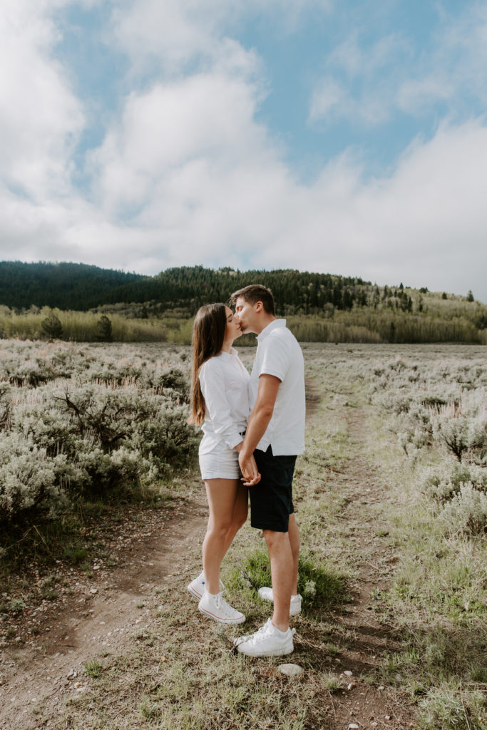 A couple standing in a field facing each other as they share a kiss with the mountains behind them in Jackson Hole