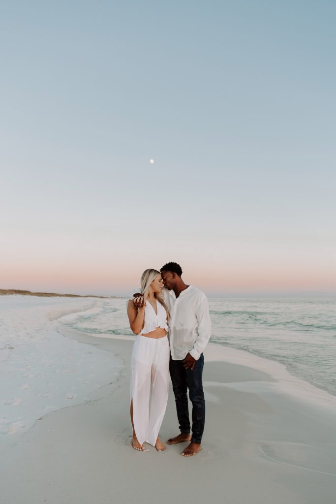 Couple standing with the man having his arm around his partners shoulders as they lean their foreheads together, the sky is painted different colors of pinks and blues with the moon shinning in the middle