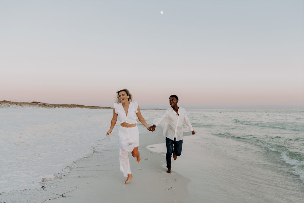 Couple holding hands and running down the beach with big smiles on their faces and the moon in the sky behind them during their beach photos in Florida