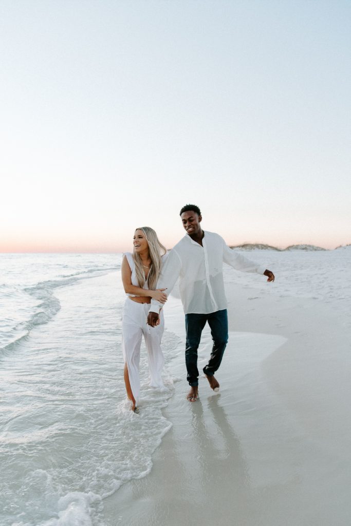 Couple holding hands and walking along the ocean as they are both smiling during their beach sunset photoshoot in Florida