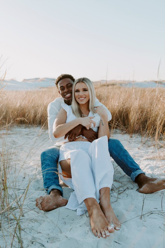 Couple sitting in each others arms in the sand, with smiels surrounded by dune grass, looking straight forward during their beach couple photos in Destin, Florida