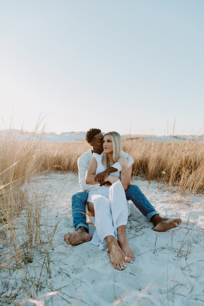 Couple sitting in a field of dune grass with the man holding his partner as she looks off into the mistaken during their beach couple photos in Destin