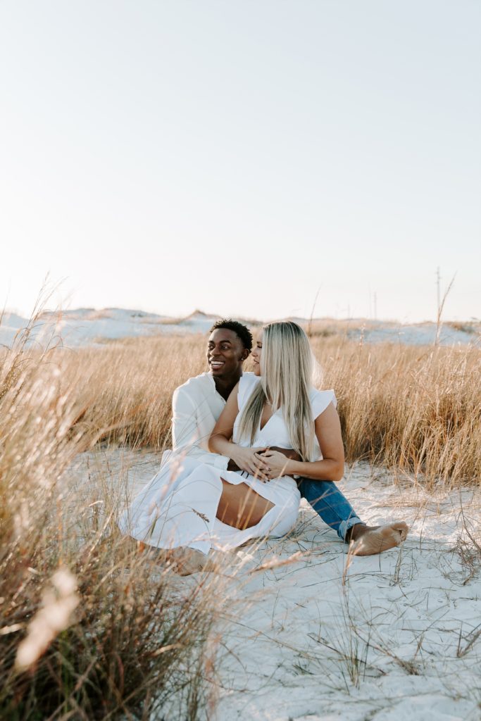 Couple sitting in the sand with the woman looking at her partner and her partner looking off laughing during their sunset beach engagement photos