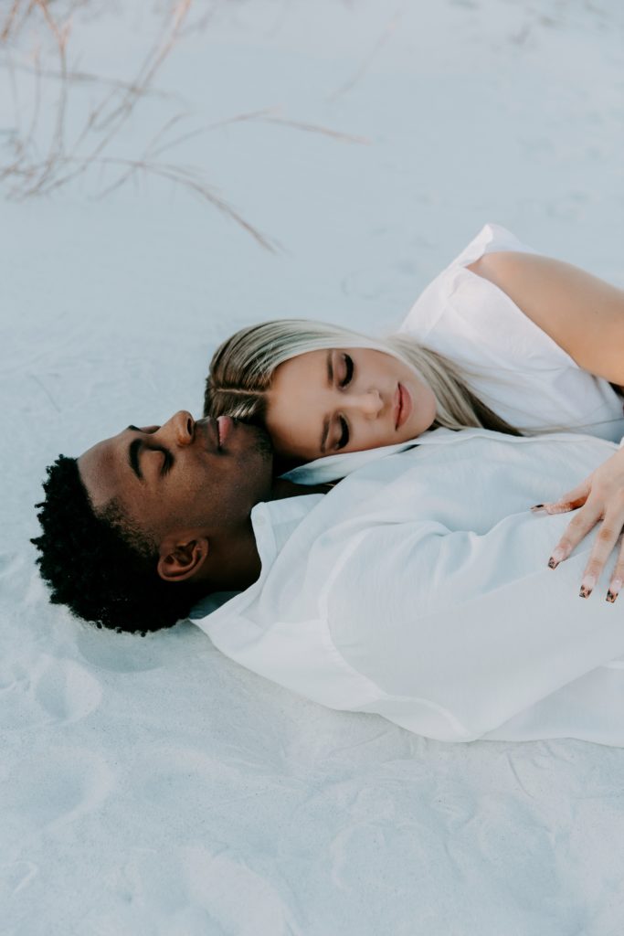 A mand and a woman laying in the sand with the woman laying her head on her partners chest both with their eyes closed during their beach engagement photos in Florida