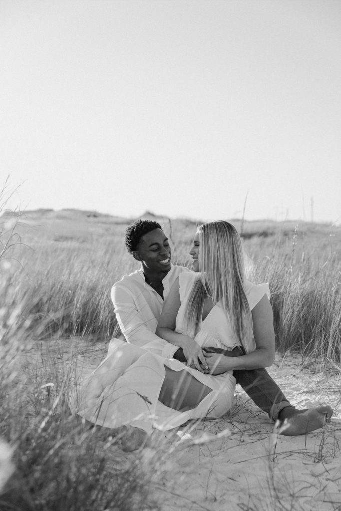 Couple sitting in the sand surrounded by beach grasses with the woman in the mans lap and they are leaning their foreheads together during their sunset engagement photos near Pensacola, FL
