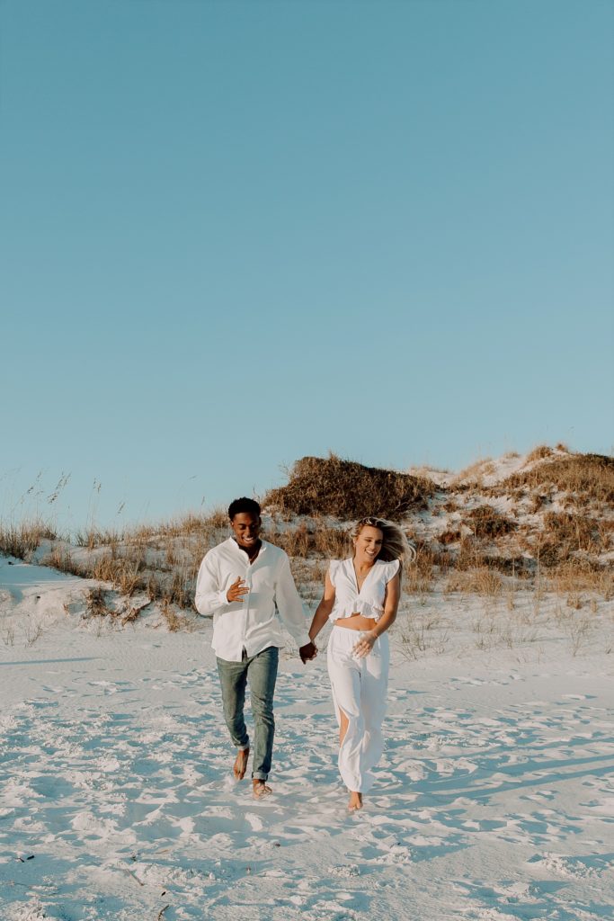 Couple holding hands and running away from the sand dunes while the womans hair sways in the wind during their winter beach engagement photos