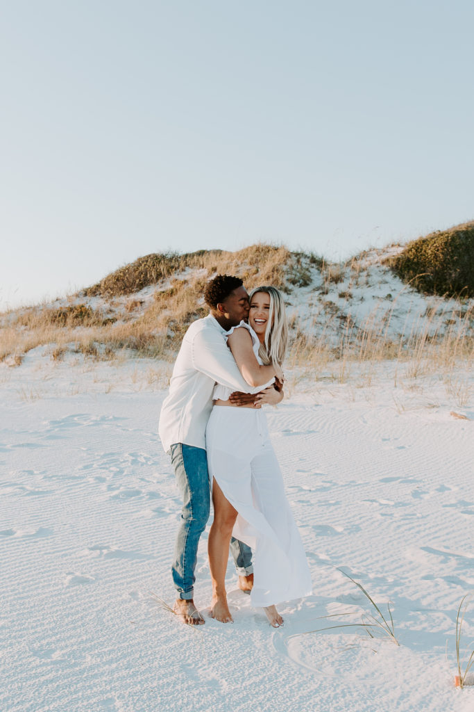 Man grabbing his partner from behind for a hug as she is laughing with sand dunes in the background during their winter beach couple photos
