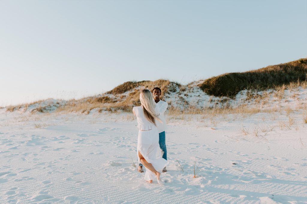 Couple swinging in circles on the beach with sand dunes in the background during their beach couple session in the Florida panhandle