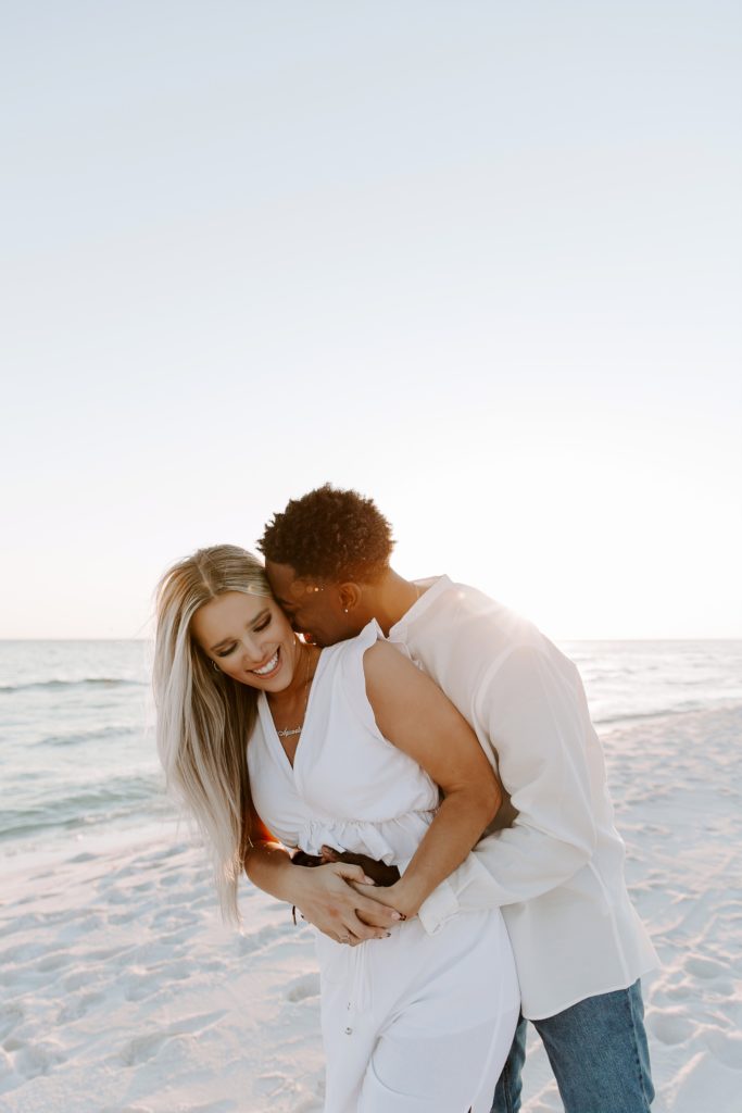 Man holding his partner from behind as he is nuzzling into her neck and holding her around the waist on the beach during their Florida couple photos
