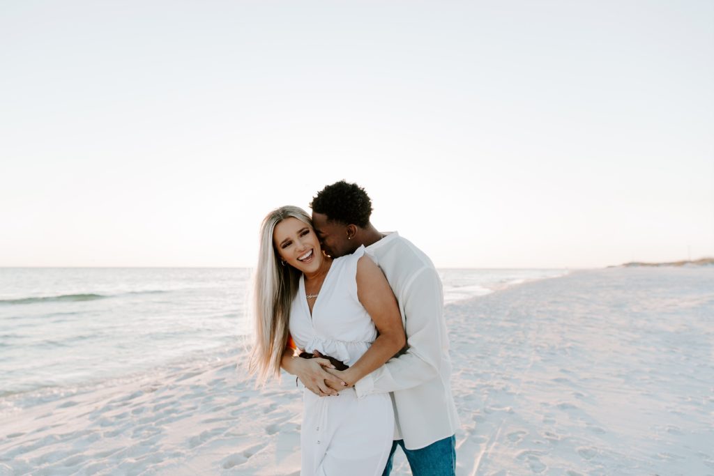 Man hugging his partner from behind as he is nuzzling into her neck and holding her around the waist on the beach during their Destin couple photos