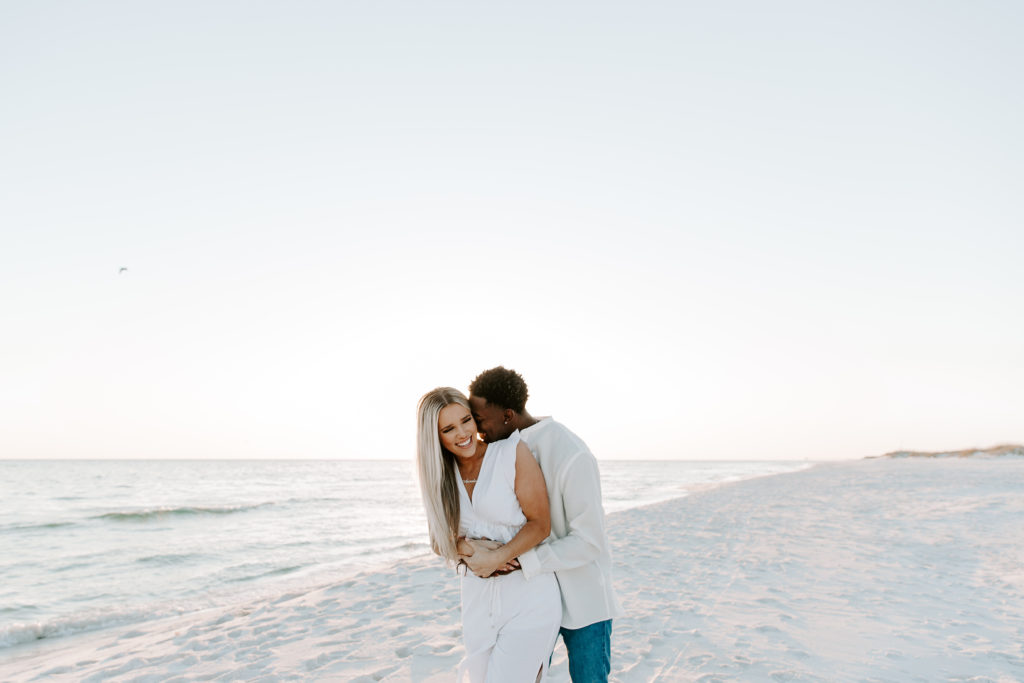 Man hugging his partner form behind as she is smiling during their beach engagement photos in Destin