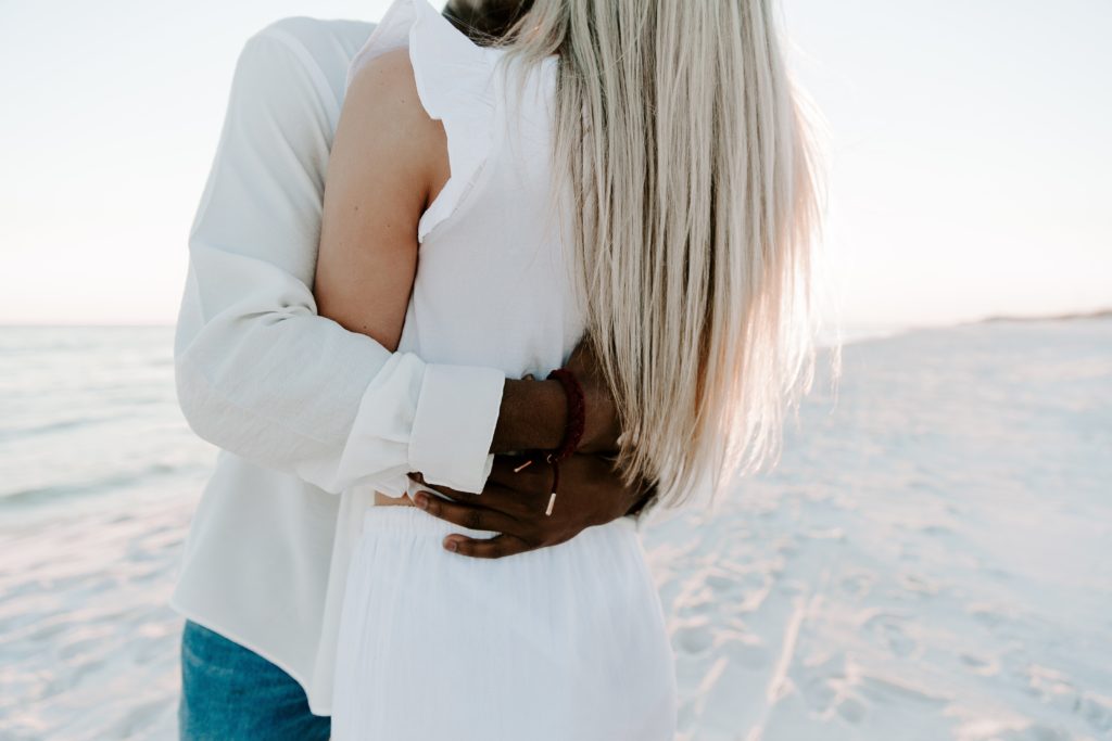 A mans arms wrapped around his partners back during their beach photoshoot in Destin, Florida