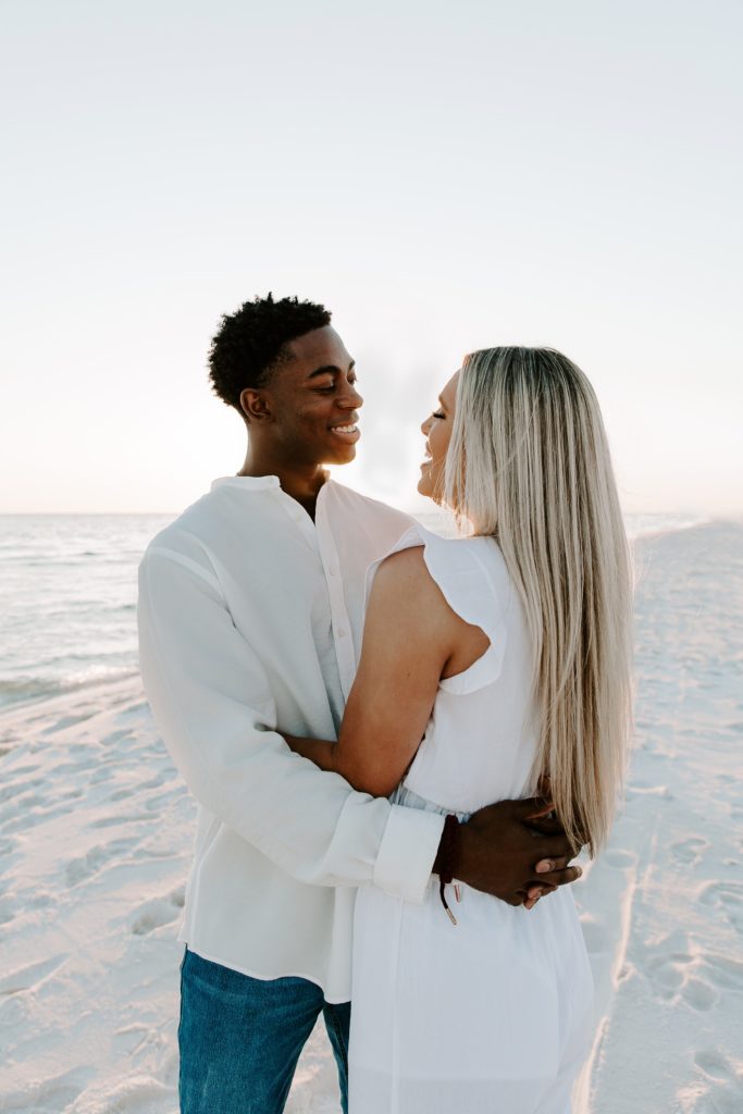 Couple standing opposite directions of each other on the beach as they are looking at each other with their arms around the other during their beach couple photos