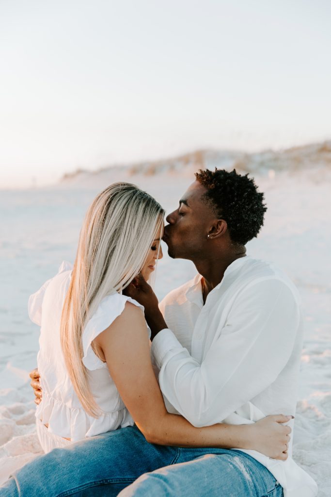 Couple sitting on the beach while the man gives his partner a kiss on the forehead during their beach couple photos